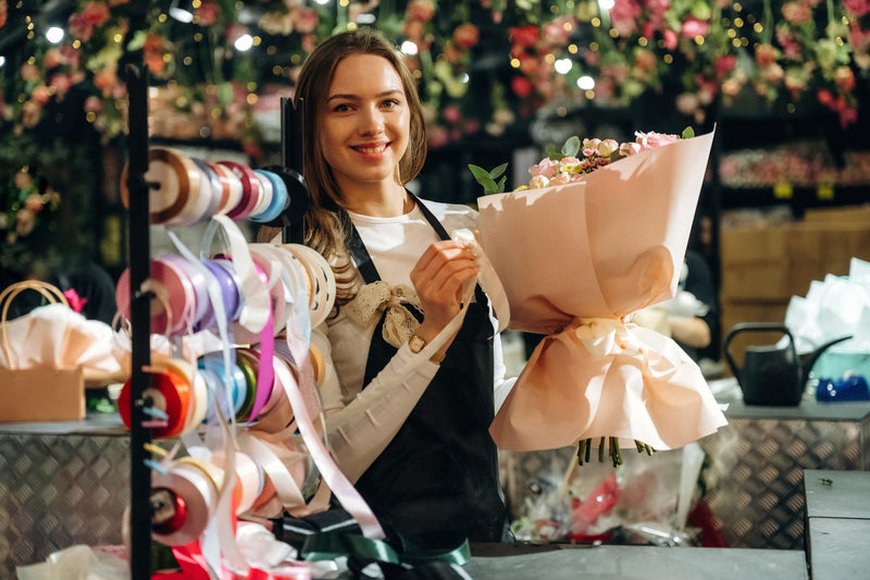 Florist holding a bouquet wrapped in flower wrapping paper by a ribbon rack